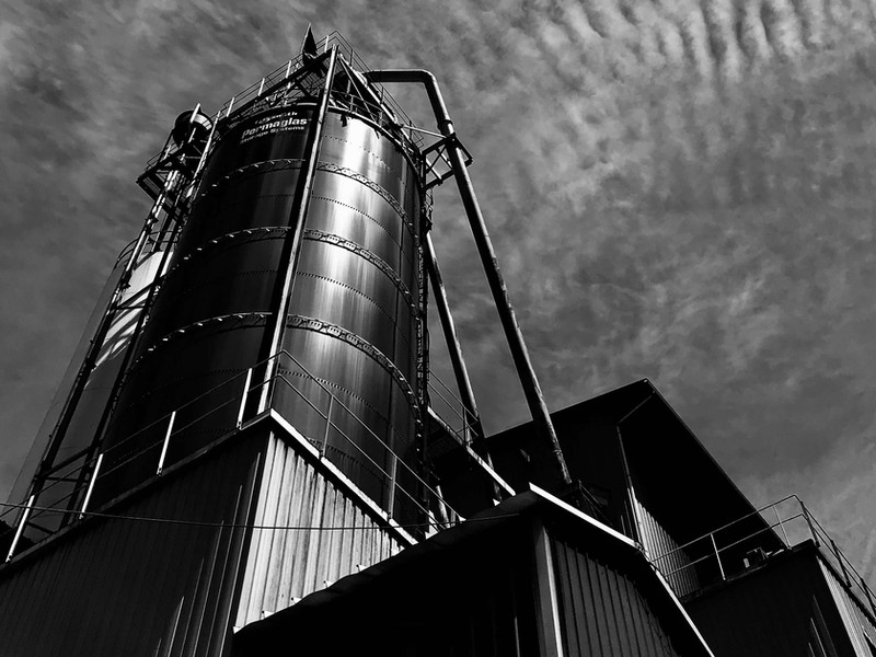 Black and white photo of an industrial silo with connected piping against a textured sky